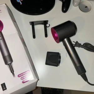 Dyson hårfön - Modern hårfön med intelligent temperaturinställning, kalluftsfunktion och inbyggt filter. Snygg design i svart och rosa, med tillbehör och originalkartong. Produkten ser oanvänd ut och är i mycket gott skick, perfekt för snabb och skonsam hårtorkning.