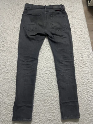 Grå skinny jeans Tommy Hilfiger - Snygga grå jeans från Tommy Hilfiger med klassisk femficksdesign och diskret logga på bakfickan. Modellen är skinny med smal passform och raka ben. Jeansen har detaljer i Hilfigers signaturfärger vid midjan. Perfekt för en stilren look. Aldrig använda (32 midja, 32 längd)