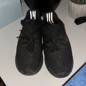 Adidas Yeezy Boost 350 V2 Black - Säljer ett par svarta Adidas Yeezy Boost 350 V2 med röd SPLY-350 text på sidan. Skorna har stickad ovandel, snörning och en ribbad platt sula med boost-teknologi. Perfekt för dig som gillar streetwear och vill ha en ikonisk sneaker i din samling. Storlek 37