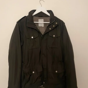 Olivgrön fieldjacket från J.Lindeberg - Fieldjacket i olivgrön från J.Lindeberg med flera fickor framtill, silverfärgade knappar och dragkedja. Jackan har hög krage, emblem på ärmen och justerbar midja. Perfekt för dig som gillar stilren och funktionell design.