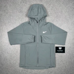 Grå vindjacka från Nike. Nike repel. - Snygg grå vindjacka från Nike med huva och hel dragkedja framtill. Jackan har lång ärm och en vit Nike-logga på bröstet. Perfekt för att hålla vinden borta under kyliga dagar. Enkel och stilren design som funkar till många outfits. Storlek M