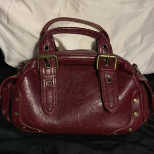 Vinröd handväska i skinn med detaljer - Snygg vinröd handväska i skinn med mässingsfärgade metalldetaljer och dragkedja upptill. Väskan har två stadiga handtag och dekorativa nitar på sidorna. Perfekt storlek för det viktigaste och ger en cool vibe till din outfit.