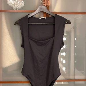 Svart ärmlös bodysuit från Zara - Snygg svart bodysuit från Zara med breda axelpartier och djup fyrkantig halsringning. Modellen är ärmlös och har en stilren, tight passform som framhäver siluetten. Perfekt att styla med jeans eller kjol för en trendig look.