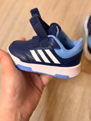 Mörkblå Adidas sneakers för barn - Säljer ett par mörkblå Adidas sneakers med vita ränder och ljusblå detaljer. Skorna har två praktiska kardborreband och en platt, vit sula med blått mönster undertill. Insidan är mjukt fodrad i ljusblått och ovandelen är i syntetmaterial.