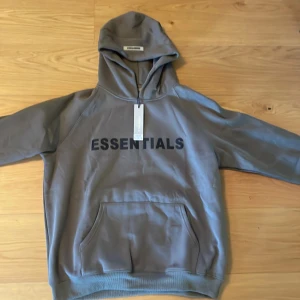 Grå/mörk grön Essentials Hoodie - Snygg grå hoodie från Essentials med svart logga framtill. Klassisk känguruficka, huva och ribbade muddar. Perfekt för en chill och avslappnad stil. Materialet känns mjukt och skönt mot huden. Hoodien är använd 1-2 gånger.