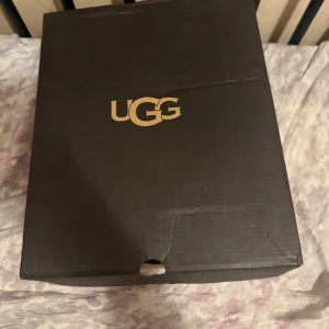 Svarta UGG boots med mönstrade snören - Säljer ett par svarta UGG boots med stickad ovandel och fluffigt foder. Skorna har mönstrade snören i svart och grått, rund tå och platt sula. Perfekta för kalla dagar och levereras i originalkartong med extra skosnören.