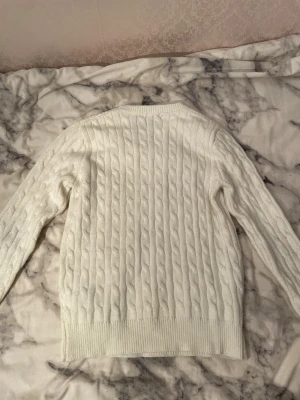 Vit kabelstickad tröja Polo Ralph Lauren - Vit kabelstickad tröja från Polo Ralph Lauren med klassisk rund hals och broderad marinblå logga på bröstet. Tröjan har långärmad passform och ribbade muddar vid ärmslut och nederkant. Perfekt för dig som gillar stilrena och tidlösa plagg.