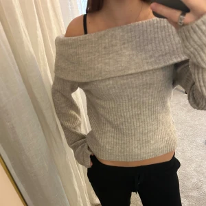 Ljusgrå/beige ribbad offshoulder stickad tröja - Superfin ljusgrå stickad tröja med ribbad struktur och bred offshoulder-krage. Tröjan har lång ärm och sitter snyggt med en slim passform. Perfekt för dig som gillar en chill men ändå stilren look.
