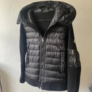 Svart Moncler Cardigan - Svart cardigan från Moncler med stickade ärmar och huva. Jackan har tvåvägsdragkedja, fickor med dragkedja på ärmen och quiltad front. Klassisk Moncler-logga på vänster ärm. Perfekt för dig som vill ha en snygg och varm jacka med street vibe.
