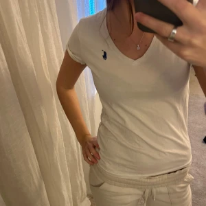 Vit t-shirt från Ralph Lauren - Vit t-shirt från Ralph Lauren med blå logga broderad på bröstet. Modellen har v-ringad hals och korta ärmar, perfekt för en clean och enkel stil. Materialet är mjuk bomull som känns skönt mot huden. Passformen är normal och t-shirten är lätt att matcha med allt. (Pytte liten fläck under bröstet! Men inget märkvärdigt)