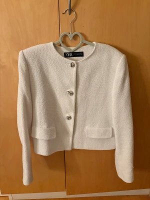 Vit kavajjacka från Zara, storlek L - Stilren vit kavajjacka från Zara i storlek L. Jackan har en rak, croppad passform, rund halsringning och snygga silverfärgade knappar framtill. Tillverkad i ett strukturerat tyg med två fickor framtill. Perfekt för en clean och trendig look.