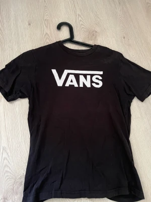 Svart Vans t-shirt med logga - Svart t-shirt från Vans med klassisk vit logga tryckt på bröstet. Modellen är kortärmad och har en rund halsringning. Tillverkad i 100% bomull för skön och avslappnad känsla. Perfekt för dig som gillar streetwear och enkel stil.