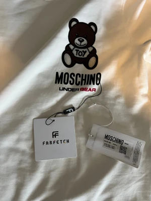 Vit Moschino sweatshirt - Snygg vit sweatshirt från Moschino med ett stort tryck av den ikoniska bruna björnen och texten 'Toy' på bröstet. Modellen har rund hals och långa ärmar. Materialet är mjuk bomull och passformen är normal. Perfekt statement-plagg för dig som gillar designer och lekfull stil.