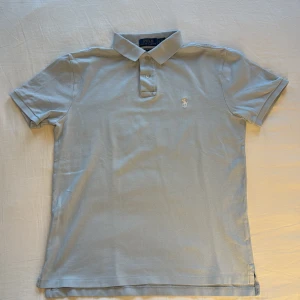 Ljusblå piké från Polo Ralph Lauren - Snygg ljusgrå pikétröja från Polo Ralph Lauren med klassisk krage och två knappar framtill. Custom slim fit-modell med broderad logga på bröstet. Tillverkad i mjuk bomull som är skön mot huden. Perfekt för en clean och stilren look. Priset går att diskuteras vid snabbaffär!