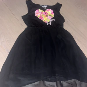 Svart klänning med blommigt hjärta Guess Barn klänning  - Supersöt svart klänning från Guess Kids med ett färgglatt blommigt hjärta mitt på bröstkorgen. Ärmlös modell med markerad midja och lätt utställd kjol i tunt material. Storleken är för 5-6 åriga barn.