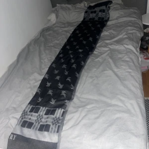 Burberry halsduk med rutor och logga - Supersnygg och lång halsduk från Burberry i grått och svart. Den har klassiskt rutmönster i ändarna och Burberrys ikoniska ryttare tryckt över hela halsduken. Perfekt accessoar för att lyfta din outfit med lite lyxig vibe.Halsduken är 2 Meter lång men går såklart att vika. 2 sidor, en svart, en grå. Har ett litet hål som inte syns!Kom med förslag