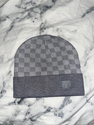 Grå rutig mössa från Louis Vuitton - Snygg grå mössa från Louis Vuitton med klassiskt rutigt mönster och vikt kant. LV-logga framtill i matchande grått. Tillverkad i mjukt stickat material som känns lyxigt och passar perfekt när det är kallt ute.