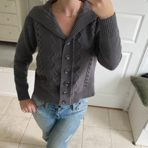 Mörkgrå stickad kofta med krage - Säljer en mörkgrå stickad kofta med bred krage och snygga kabelstickade detaljer framtill. Koftan har långa ärmar, ribbade muddar och knäppning med fem knappar. Perfekt att slänga på över en t-shirt för en chill look. Från Salvo green