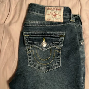True Religion jeans, Becca mid rise  - Säljer ett par blå jeans från True Religion med tydliga kontrastsömmar i gult och vitt. Klassisk femficksmodell med lock på bakfickorna och silverfärgade knappar. Snygg tvättad effekt och ikonisk logga på bakfickan. Perfekta för dig som gillar streetwear.