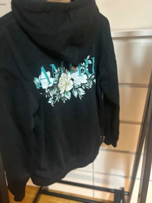 Svart hoodie med blommor från Amiri - Svart hoodie från Amiri i storlek S med blommigt tryck och logga i turkos på bröstet och ryggen. Tröjan har huva med snörning, känguruficka och är tillverkad i mjuk bomull med lite elastan för extra komfort. Perfekt för dig som gillar streetwear med stil.
