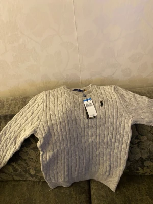 Ljusgrå stickad tröja Polo Ralph Lauren - Snygg ljusgrå stickad tröja från Polo Ralph Lauren med klassiskt kabelstickat mönster och rund hals. Tröjan har lång ärm och den ikoniska broderade loggan på bröstet. Perfekt för dig som gillar stilrena och tidlösa plagg. Pris kan diskuteras 