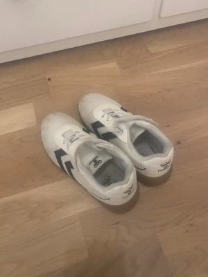 Vita Hummel sneakers strl 35 - Säljer ett par vita Hummel sneakers med svarta detaljer och gummisula. Skorna har klassisk snörning och är tillverkade i mesh och syntetmaterial för bra ventilation. Snygg sportig design med Hummels ikoniska pilar på sidan. Perfekt för dig som gillar stilrena och bekväma skor.