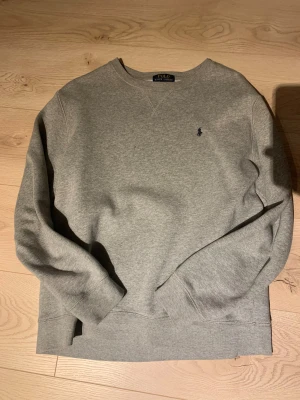 Grå sweatshirt från Polo Ralph Lauren - Säljer en stilren grå sweatshirt från Polo Ralph Lauren med klassisk rund hals och långärmad design. Tröjan har ett litet mörkblått Polo-logo broderat på bröstet och är gjord i mjuk bomullsmix med fleece på insidan för extra komfort.