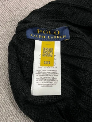 Ralph Lauren Knitwear Mössa - Perfekt nu när snön har fallit! Svart knitwear mössa från Polo Ralph Lauren med klassisk ribbad kant och vit broderad logga framtill. Tillverkad i 100% bomull för skön komfort och stilren look. Perfekt accessoar för att lyfta din outfit under kyliga dagar.