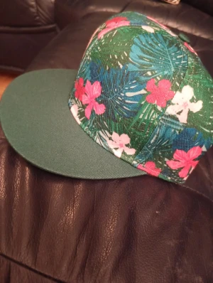 Grön keps med blommigt mönster - Snygg keps med grön skärm och blommigt mönster i rosa, vitt och blått på ovansidan. Perfekt för dig som vill sticka ut med färg och tropisk vibe. Kepsen har platt skärm och är tillverkad i bomullsmaterial.