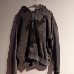 Grå hoodie med tryck från Bershka - Säljer en mörkgrå hoodie från Bershka i storlek L. Tröjan har huva, snörning och dragkedja framtill samt ett coolt tryck på bröstet. Materialet är mjukt och känns skönt mot huden, perfekt för höst/vår dagar. Långa ärmar och ribbade muddar ger en avslappnad vibe.