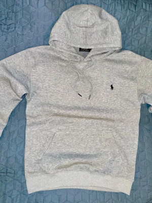 Grå hoodie från Polo Ralph Lauren - Snygg ljusgrå hoodie från Polo Ralph Lauren med klassisk huva och dragsnören. Broderad logga i svart på bröstet och stor känguruficka framtill. 
