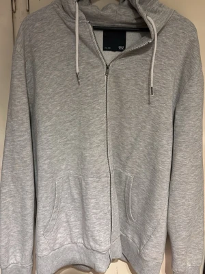 Ljusgrå zip hoodie från 157 - Ljusgrå hoodie med dragkedja från 157. Klassisk modell med huva och vita dragsnören, samt två fickor framtill. Tillverkad i mjukt sweatshirtmaterial som är perfekt för chill dagar. Enkel och stilren design som passar till det mesta. Väldigt fint skick på utsidan, dock lite uttöjd, så passar nog både XL-XXL