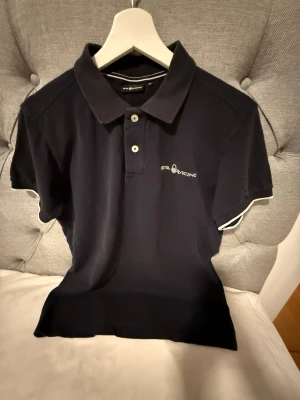 Marinblå pikétröja från Sail Racing - Klassisk marinblå pikétröja från Sail Racing med vit logga på bröstet. Tröjan har krage, två knappar framtill och korta ärmar med vit detalj vid ärmslut. Perfekt till en avslappnad men stilren look.