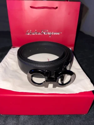 Svart skärp från Ferragamo - Snyggt svart skärp från Salvatore Ferragamo med ikoniskt dubbel-Gancini-spänne i silverfärg. Bältet är gjort i slätt skinn och har en stilren, modern look som passar till många outfits.