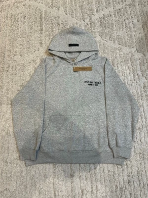 Grå hoodie från Essentials Fear of God - Snygg grå hoodie från Essentials Fear of God med svart logga på bröstet. Klassisk passform med huva och känguruficka framtill. Tillverkad i mjukt bomullsmaterial som är perfekt för en avslappnad stil.