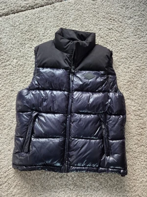 Svart/blå dunväst från replay - Snygg svart dunväst från replay med glansig finish och hög krage. Västen har två dragkedjefickor framtill och en diskret logga på bröstet. Över axlarna och kragen är materialet mattare för en cool kontrast. Skriv vid frågor.