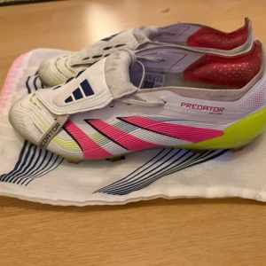 Adidas Predator fotbollsskor vit/röd - Säljer ett par Adidas Predator fotbollsskor i vitt med röda och neon-gula detaljer. Skorna har klassisk Predator-design med svarta ränder och text, samt låg profil och snörning. Perfekta för dig som vill sticka ut på planen. Felfria förutom en defekt (se bild 5) köpta i december och passar inte mig längre hör av dig vid frågor påse ingår!😁🤩