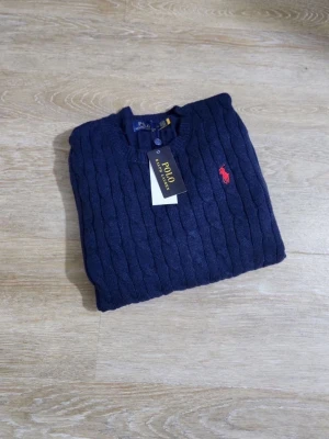 Mörkblå kabelstickad tröja Polo Ralph Lauren - Mörkblå kabelstickad tröja från Polo Ralph Lauren med rund halsringning och klassisk röd broderad logga på bröstet. Tröjan har ribbade muddar och är tillverkad i mjukt material som ger en stilren och tidlös look.