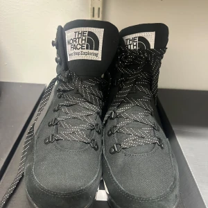 Svarta kängor från The North Face 42 - Snygga svarta kängor från The North Face med högt skaft och robust snörning. Kängorna har vit logotyp på plösen och sidan, samt svarta och vita mönstrade skosnören. Ovandelen är i slitstark textil och sulan är vit med svart yttersula.