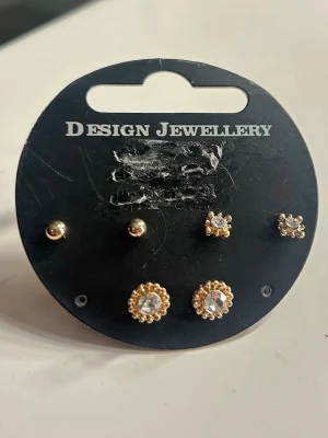 Guldiga örhängen 3 par - Säljer ett set med tre par guldiga örhängen från Design Jewellery. Två par har gnistrande klara stenar, varav ett par är omgivna av små guldfärgade pärlor. Aldrig använda.