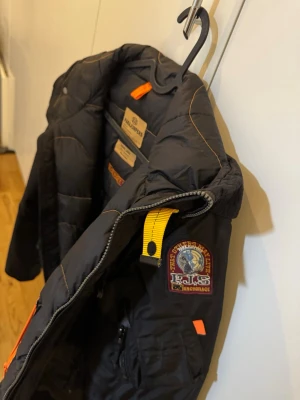 Svart dunjacka från Parajumpers - Säljer en svart dunjacka från Parajumpers med gul detalj vid kragen och patch på ärmen. Jackan har dragkedja, flera fickor och orangea detaljer. Perfekt för kalla vinterdagar och har en sportig, modern look.