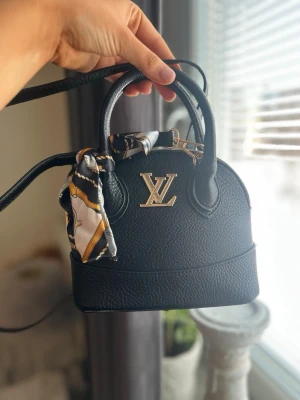 Louis Vuitton svart handväska - Snygg svart handväska från Louis Vuitton i klassisk modell med guldfärgad LV-logga framtill. Väskan är i strukturerat läder och har både handtag och axelrem. Dekorerad med en scarf i svart, vitt och guld. Perfekt accessoar för en trendig look. PRIS KAN DISKUTERAS 