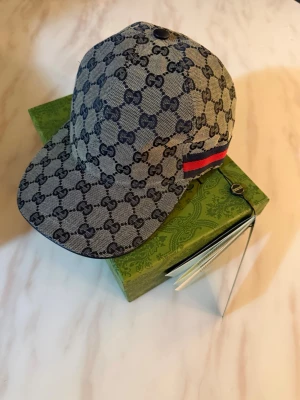 Gucci keps med GG-mönster - Snygg keps från Gucci i beige canvas med klassiskt GG-monogram över hela kepsen. Röd och blå rand på sidan ger en extra touch. Justerbar baktill och med böjd skärm. Perfekt accessoar för dig som gillar lyxiga detaljer.