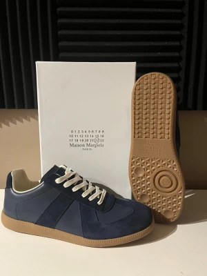 Maison Margiela blå sneakers -  Hej säljer ett par fett snygga skor i str 45 men passar också 44. Snygga blå sneakers från Maison Margiela med ovandel i mocka och läder, vita skosnören och klassisk gummisula i beige. Ikonisk design med diskreta detaljer och låg silhuett som passar perfekt till streetwear och casual outfits.