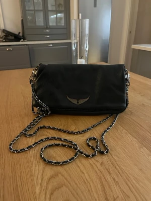 Svart axelväska från Zadig & Voltaire - Cool svart axelväska i mjukt läder från Zadig & Voltaire med silverfärgad kedjerem och ikonisk vinglogga framtill. Väskan har avtagbar kedja och dragkedjedetaljer. Perfekt för dig som gillar en stilren men edgy look. Väskan är äkta men väll använd och därför lågt pris!