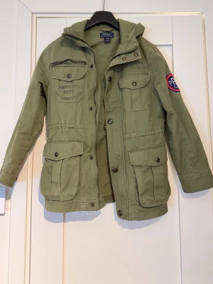 Olivgrön parkas från Polo Ralph Lauren - Det är min sons jacka, passar till 10-13år. Snygg olivgrön parkas från Polo Ralph Lauren med huva, flera stora fickor och patch på ärmen. Jackan har dragkedja och knappar framtill samt coola detaljer som tryck på bröstet. Perfekt för dig som gillar utility-stil och streetwear.