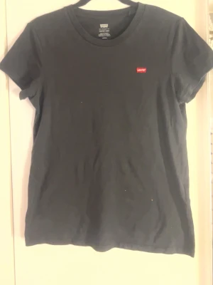 Svart t-shirt från Levi's - Klassisk svart t-shirt från Levi's med rund halsringning och liten röd logotyp på bröstet. Enkel och stilren design som passar till det mesta. Tillverkad i mjuk bomull och har korta ärmar.