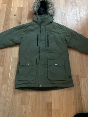 Olivgrön parkas med fuskpäls - Snygg olivgrön parkas med huva och fuskpäls, två stora fickor med lock och tryckknappar samt två bröstfickor med dragkedja. Jackan är vadderad och har svart innerfoder. Perfekt för kalla dagar och riktigt skön att bära.