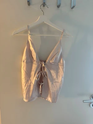 Ljusrosa satinlinne med rosett - Snyggt ljusrosa linne i satin med smala axelband och dekorativ rosett framtill. Linnet har en djup v-ringning och en mjuk, glansig finish som ger en lyxig känsla. I storlek S