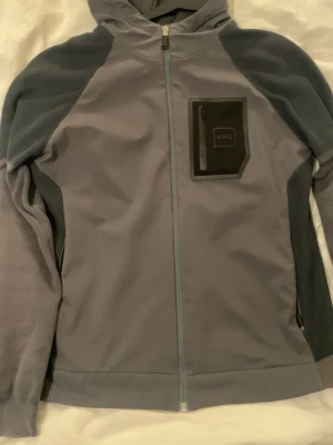 Grå hoodie från BOSS med dragkedja - Snygg grå hoodie från BOSS med hel dragkedja framtill och kontrasterande mörkare partier på ärmar och sidor. Praktisk huva och en cool bröstficka med BOSS-logga. Perfekt för en avslappnad streetwear-look. Tillverkad i mjukt material för extra komfort.